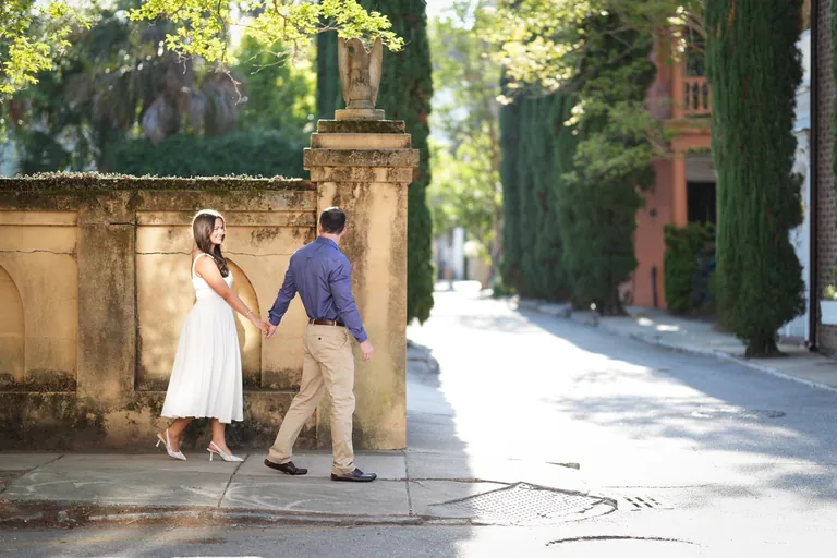 A couple walking hand in hand past an old stone wall lined with cypress trees