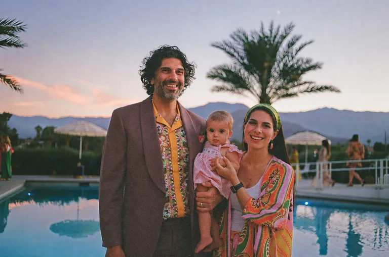 Parents holding their toddler beside a pool with palm trees behind them