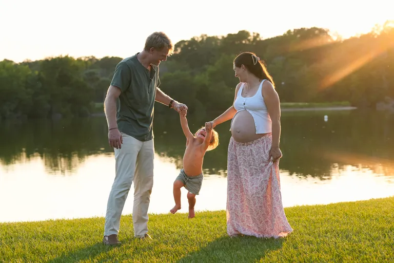 Expecting parents swinging their toddler by the hands beside the lake at golden hour