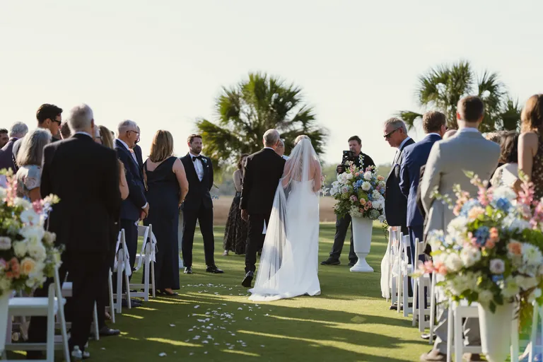 A bride walking down the aisle past standing guests and palm trees